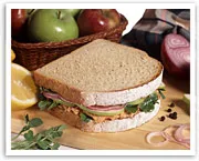 Sandwich with vegetables on cutting board, fruit basket, and sliced onions in background.