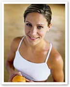 A woman in a white tank top holding an orange, smiling at the camera, suggesting a healthy lifestyle.