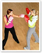 Two women practicing boxing in a gym, one wearing pink gloves throwing a punch while the other holds pads for training.
