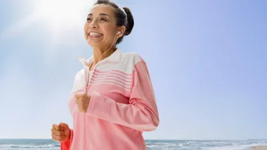 A woman in a pink tracksuit jogs on the beach with earbuds in, smiling under a clear blue sky.