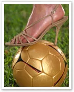 A foot in a high-heeled sandal rests on a gold soccer ball on grass.