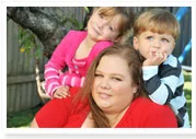 A woman in a red shirt with two children leaning on her shoulders, outdoors in a garden setting.