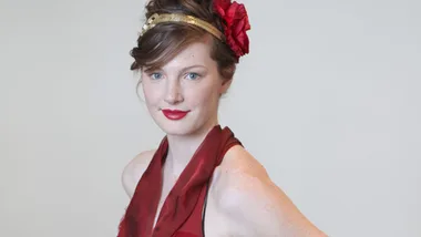 A woman with a red dress and floral headpiece smiles, set against a light gray background.