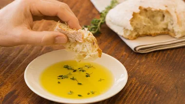 A hand dips bread into a small bowl of olive oil with herbs, placed on a wooden table next to more bread and a napkin.