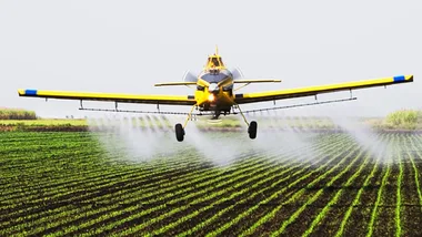Crop-dusting yellow airplane spraying pesticides over green farmland.