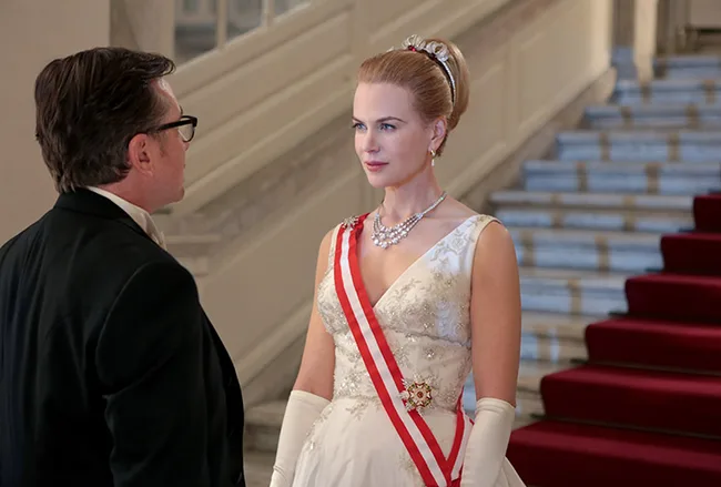 Nicole Kidman as Grace of Monaco in a formal gown, adorned with a red sash and pearl necklace, stands regally indoors.