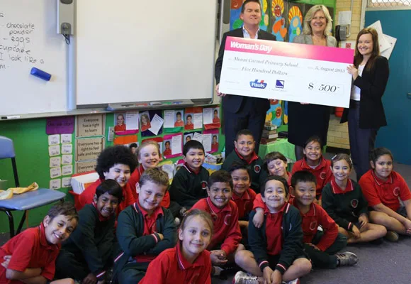 Group of adults and children smiling in a classroom with a large $500 school donation check.