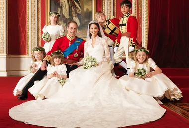 Bride and groom in royal attire, surrounded by flower girls and page boys in a formal setting.