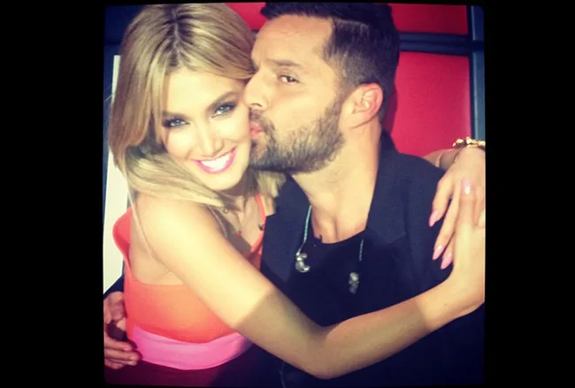 Two judges from "The Voice" taking a close, joyful selfie; one is kissing the other on the cheek.