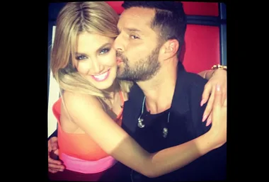 Two judges from "The Voice" taking a close, joyful selfie; one is kissing the other on the cheek.