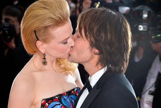 A couple kisses on the red carpet, surrounded by photographers and flashing cameras.