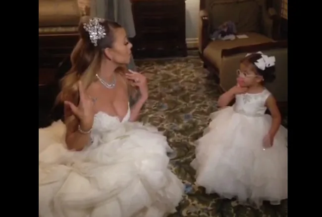 Bride in elegant gown and jeweled headpiece sits with a young girl in a white dress and glasses, both in a formal setting.