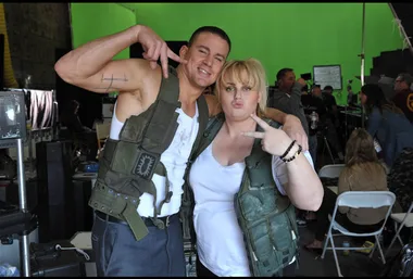 Two actors in tactical vests pose playfully on a film set with a green screen backdrop and crew in the background.