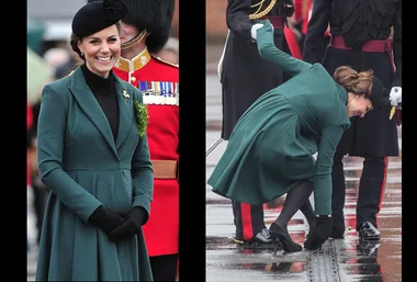 A woman in a green coat with a hat smiles and bends down in a public event, showing her dress partially lifted by the wind.