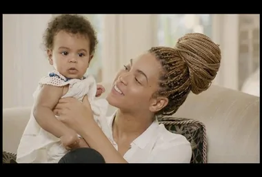 A woman with braided hair lovingly holds an infant on a sofa in a bright room.