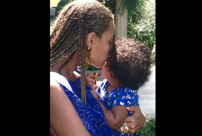 Mother in blue dress, braided hair, holds and kisses a young child in a matching blue outfit outdoors.