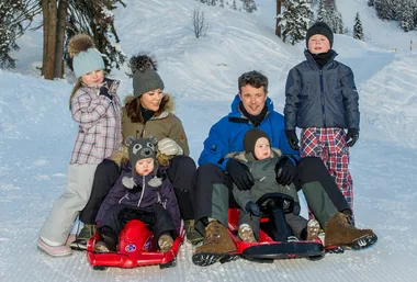 Family sledding in the snow, all dressed in winter gear, surrounded by snowy trees. Smiling and enjoying the holiday.