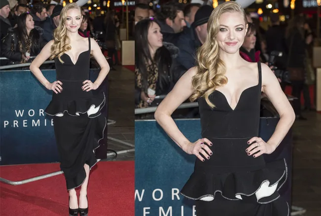 Woman in a black dress at a red carpet event, smiling with long wavy blonde hair, hands on hips, surrounded by people.