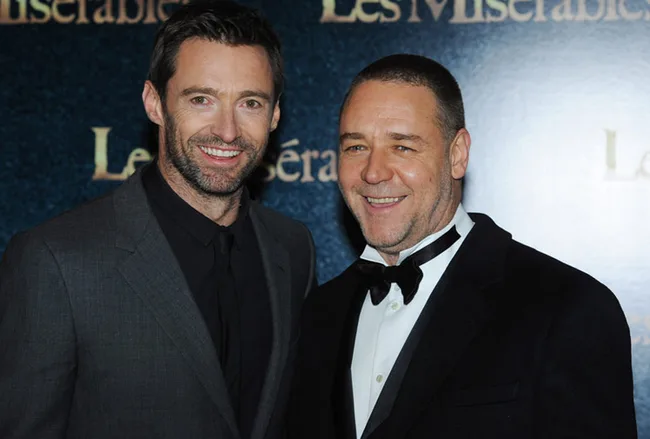 Two men in formal attire smiling at a "Les Misérables" event. One wears a black suit, the other a black tuxedo with a bow tie.