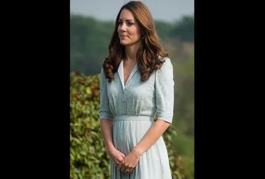 Woman standing outdoors in a light blue dress, hands clasped, with greenery in the background.