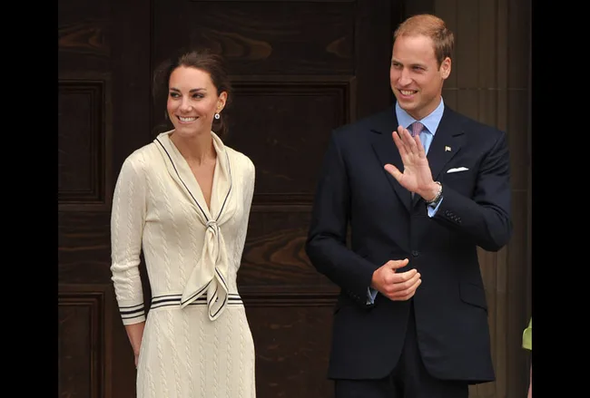 A man in a suit waves, standing beside a smiling woman in a cream dress with a black trim, both in front of a wooden door.