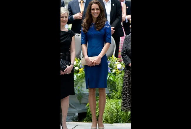 A woman in a royal blue dress stands smiling outdoors, surrounded by other people, with green plants in the background.