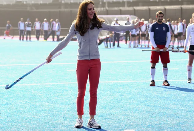 A woman in casual sportswear on a hockey field, holding a hockey stick, with people in the background.