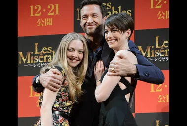 Cast members of "Les Misérables" pose together at a promotional event, smiling in front of a red and black backdrop.