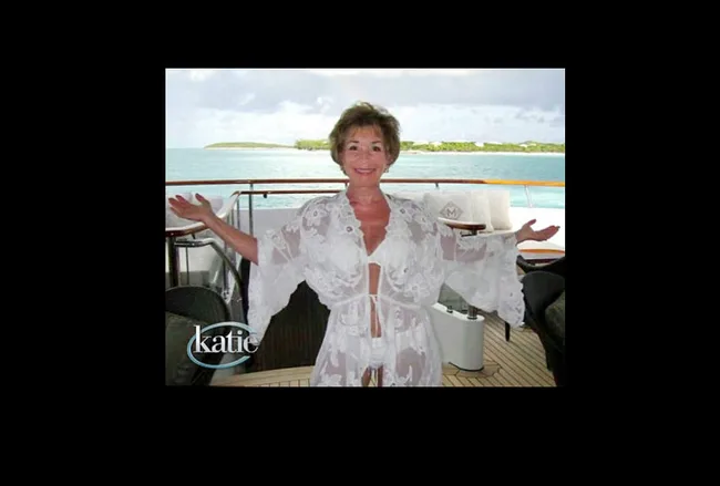 Judge Judy in a white cover-up stands on a boat, arms open wide with a scenic view of water and land in the background.