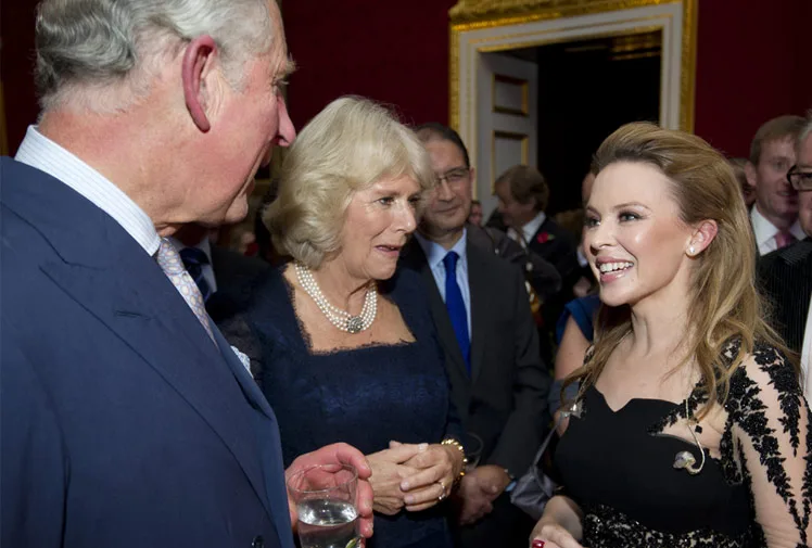 Three people at an event, including a woman in a black dress, talking animatedly in an elegant room.