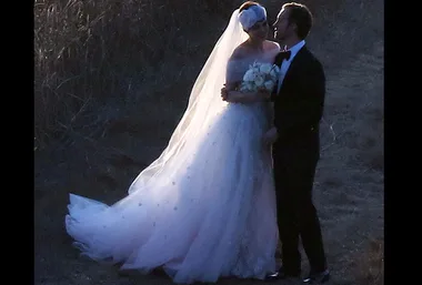 Bride in white gown and veil holding flowers, standing with groom in tuxedo, outdoors at twilight.