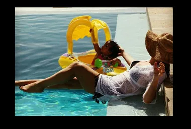 Woman in a sunhat lounges by a pool while a child plays nearby with a yellow inflatable toy.
