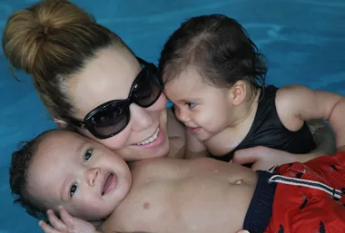 Women in sunglasses smiling with two babies in a pool, sharing a joyful moment.