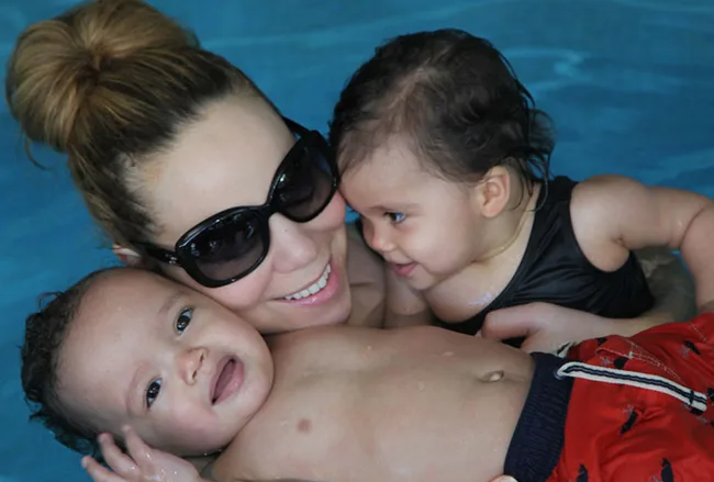 Women in sunglasses smiling with two babies in a pool, sharing a joyful moment.