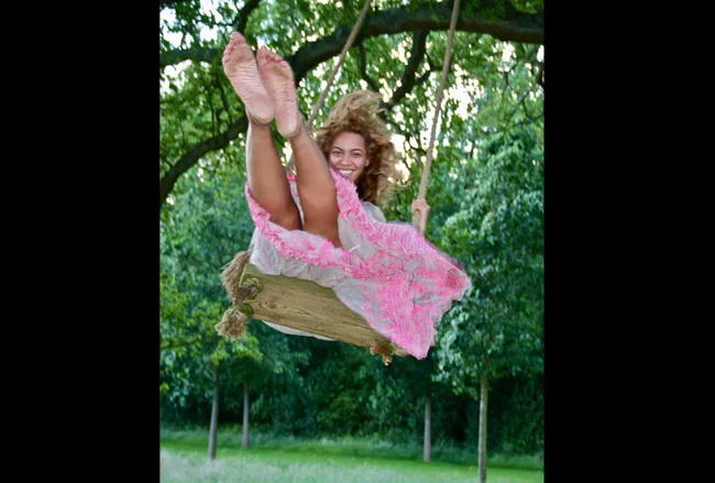 Woman in a pink dress joyfully swings on a wooden swing in a lush green garden.