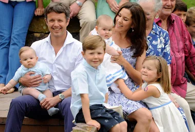 Family group photo with adults holding four children, sitting outdoors on steps, everyone smiling and posing casually.
