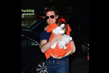 Tom Cruise carrying a child wrapped in an orange blanket with a stuffed elephant toy on a city street at night.