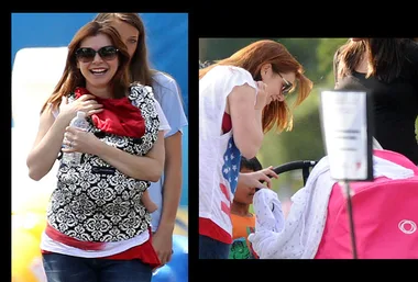 Mum with red hair, sunglasses, carries baby in sling and stroller outdoors.