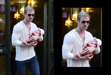 Man in sunglasses holding a baby in a red outfit and white hat, standing outside a building with glass doors.
