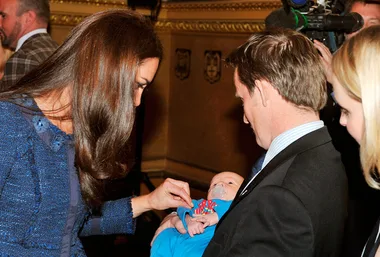 Kate Middleton admiring a baby held by a man in a suit, standing in an elegant room with cameras capturing the moment.
