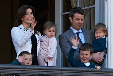 Royal family waving from a balcony, including three young children in formal attire with their parents.