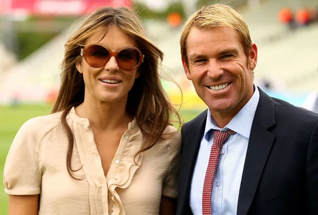 A smiling woman in sunglasses and a ruffled blouse stands next to a smiling man in a suit and tie at an outdoor event.