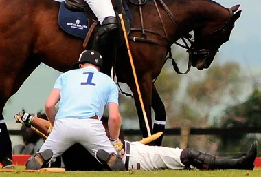 Polo player in a blue jersey tends to a fallen player on the ground with a nearby horse during a match.