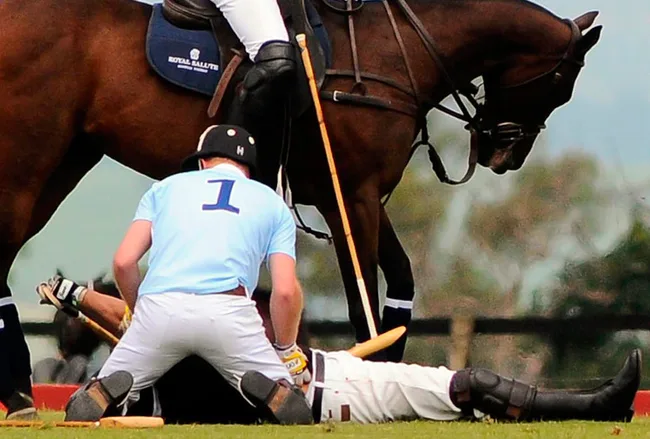 Polo player in a blue jersey tends to a fallen player on the ground with a nearby horse during a match.