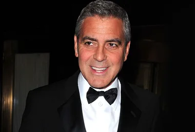 A man in a black tuxedo and bow tie, smiling at an event with a dark background.