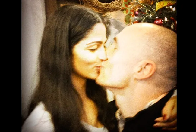 A couple sharing a kiss in a warmly lit setting with holiday decorations in the background.