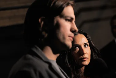 A woman looks at a man speaking at an event, with microphones visible in dim lighting.