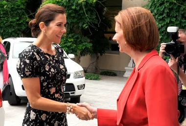Two women smiling and shaking hands outdoors, one in a floral dress, the other in a red suit, with photographers nearby.