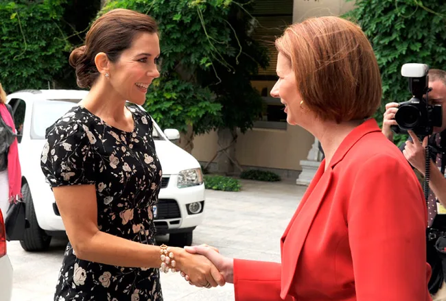 Two women smiling and shaking hands outdoors, one in a floral dress, the other in a red suit, with photographers nearby.