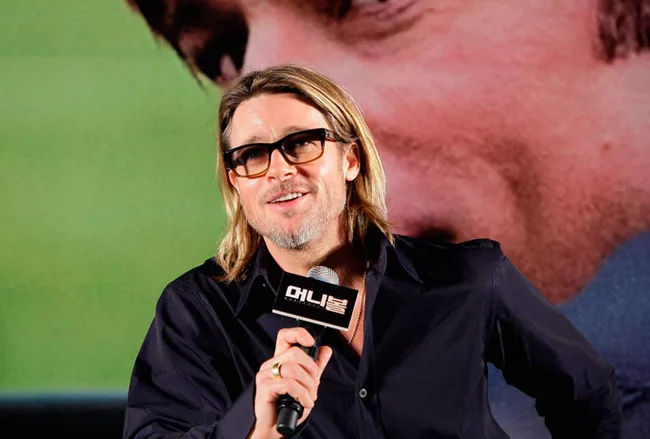A man with glasses and long hair speaks into a microphone at a public event, smiling.